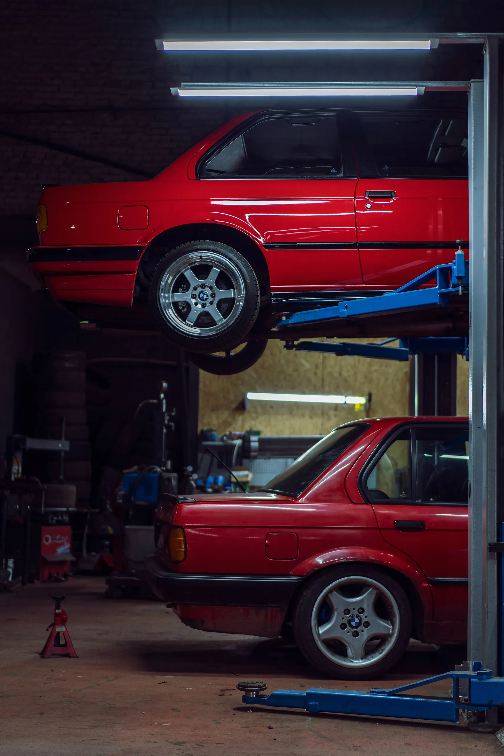 Dwa czerwone BMW E30, jedno na podnośniku w warsztacie podczas naprawy